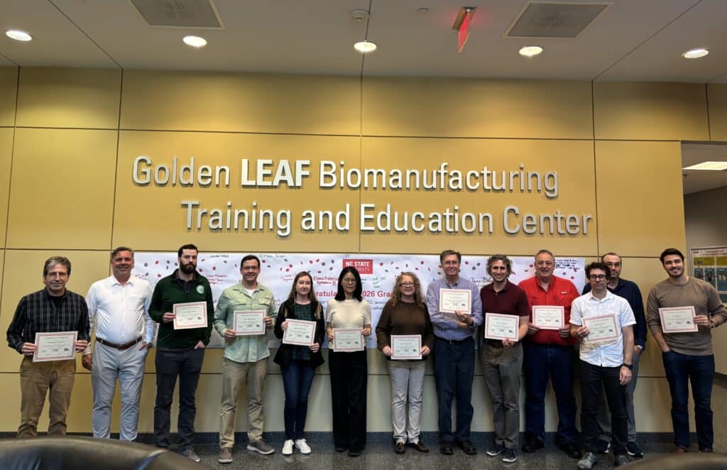 Participants of the Artificial Intelligence in FDA Regulated Manufacturing: From Foundations to Implementation course hold their Certificates of Completion in front of a wall with which reads "Golden LEAF Biomanufacturing Training and Education Center". 